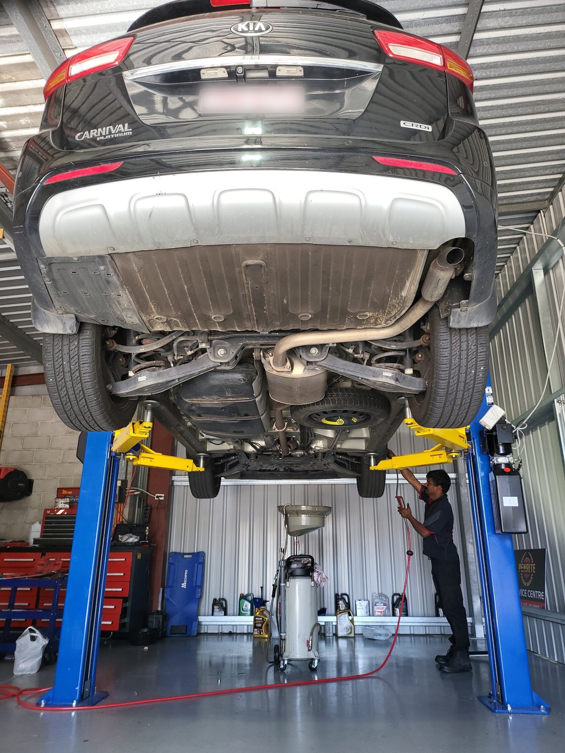 Kia Carnival raised on the hoist during a service, with the oil drain rig underneath and the mechanic checking the rear